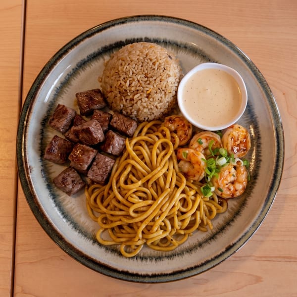Hibachi steak, shrimp, fried rice, and noodles with a side of sauce on a plate.